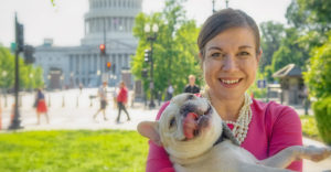 Photo of a smiling woman holding a dog in front of the U.S. Capitol Building.