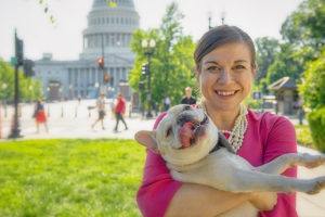 Photo of a smiling woman holding a dog in front of the U.S. Capitol Building.